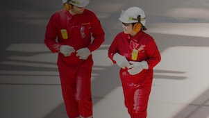 Male and female worker in red overalls and white helmets and hearing protection walking
