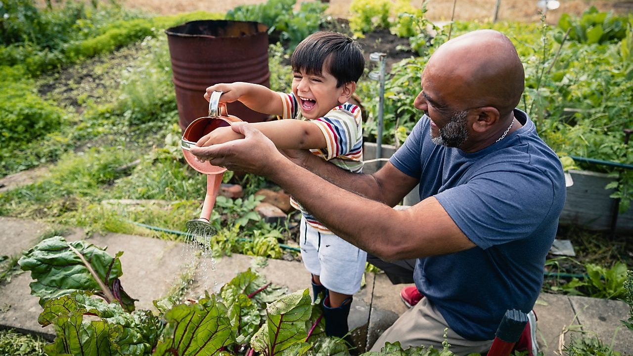 Man helping a smiling child water plants in a garden
