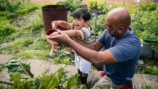 Man helping a smiling child water plants in a garden