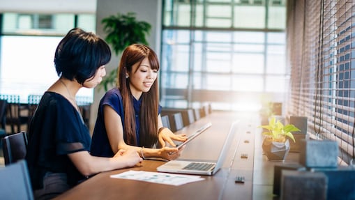 Two women sitting at a desk looking at a computer and a tablet