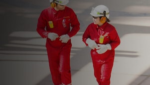 Male and female worker in red overalls and white helmets and hearing protection walking