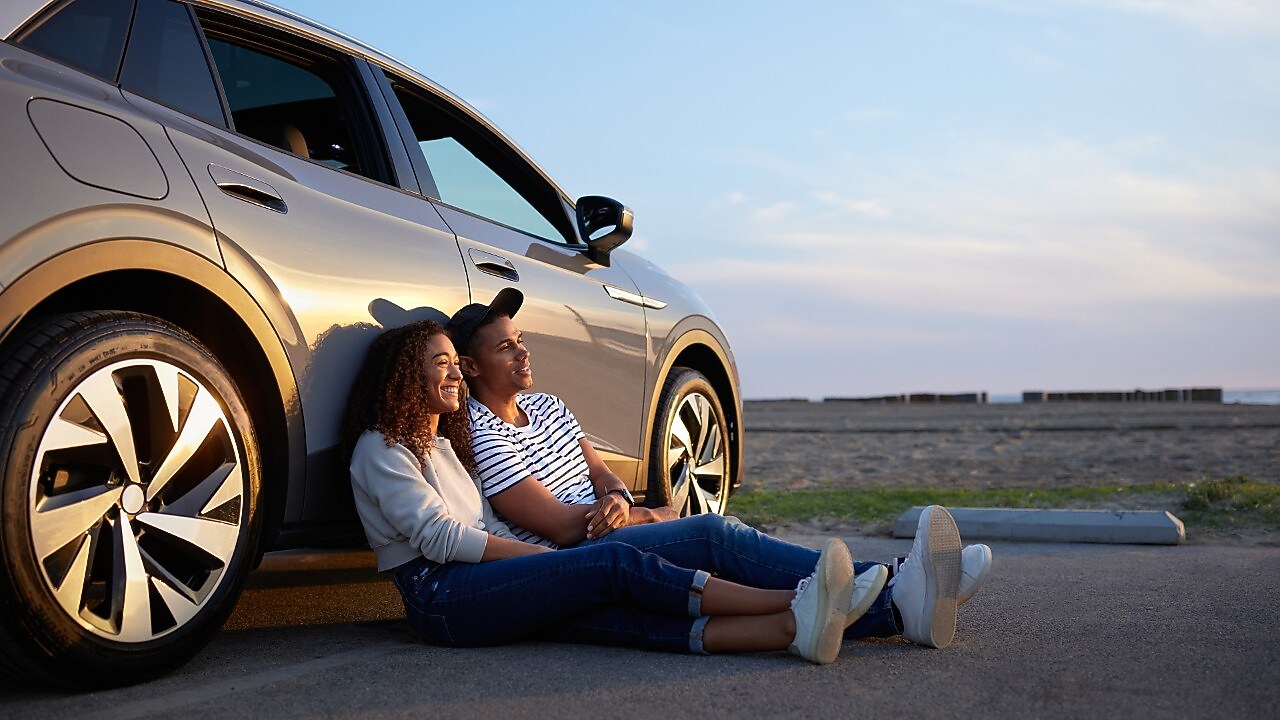 Pareja sentada frente a un auto al atardecer.