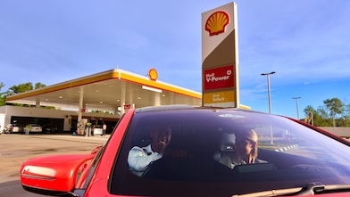 Auto rojo con dos personas frente a una estación de gasolina Shell.