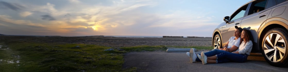 Pareja sentada frente a un auto al atardecer.