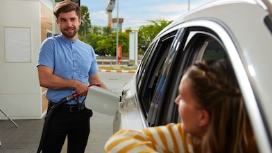Hombre llenando el tanque de gasolina mientras una mujer lo observa desde el auto.