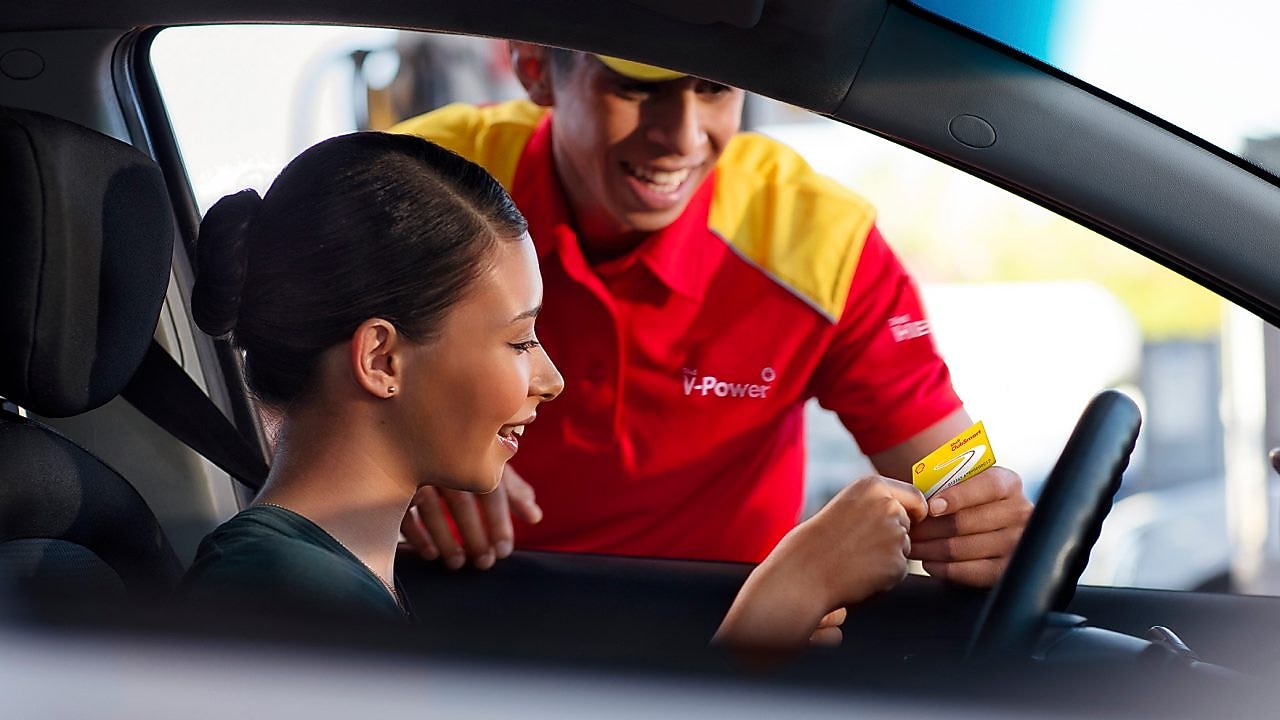 Mujer al volante, en un coche parado, siendo atendida por un empleado de una gasolinera Shell. Tiene el pelo negro y sonríe, lleva uniforme y una gorra roja. Ella le enseña una tarjeta Shell.