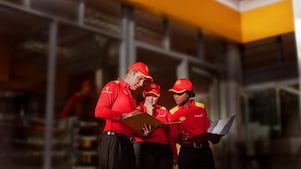 Tres empleados (un hombre y dos mujeres) con uniforme de Shell -camisa roja y gorra- examinan una carpeta que sostiene el hombre. Una de las mujeres también lleva en la mano un maletín con documentos.